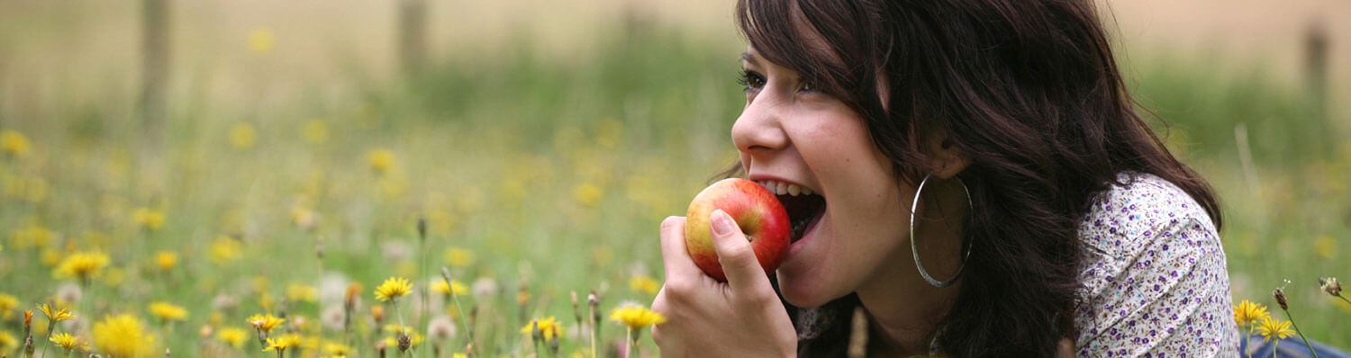 A woman with long dark hair is biting into a red apple while lying in a field of green grass and yellow flowers.