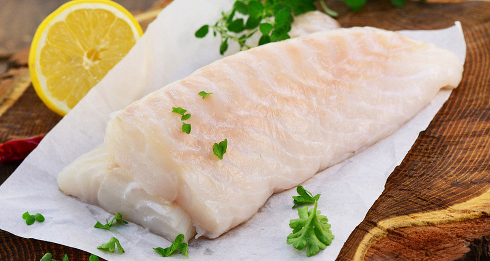 A fish fillet rests on a wooden surface, accompanied by a lemon wedge and fresh herbs.