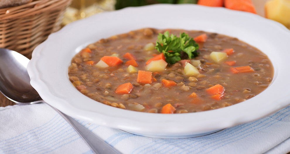 A bowl of thick, hearty soup with visible chunks of carrot, potato, and lentils is presented on a table with a spoon and a basket of bread in the background.
